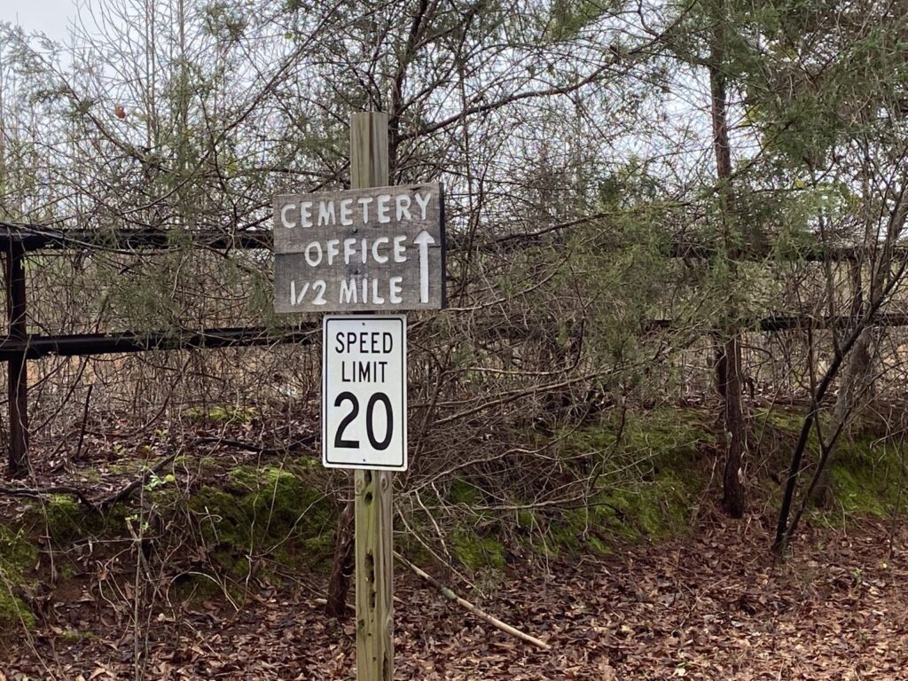 A directional sign and speed limit sign are set along a wooden fence in the woods at Honey Creek Cemetery.