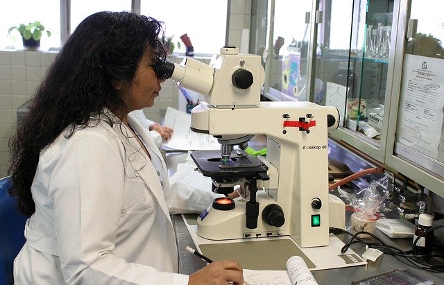 A woman in a white lab coat peers through a microscope in a laboratory.